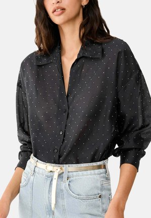 Donna che indossa una camicia nera con piccoli pois bianchi infilata in jeans azzurri chiari a vita alta con una sottile cintura beige.