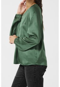 Blouse en satin vert avec un col en V, manches longues bouffantes avec poignets élastiques, et une coupe décontractée, associée à un jean foncé.