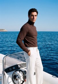 Homme portant un col roulé marron et un pantalon blanc se tient sur un bateau près du volant, avec l'océan et un ciel dégagé en arrière-plan.