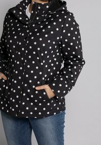 Chaqueta negra impermeable con lunares blancos, que presenta un cierre frontal con cremallera, cierres de botón y bolsillos laterales. Tejido texturizado, diseño con capucha.