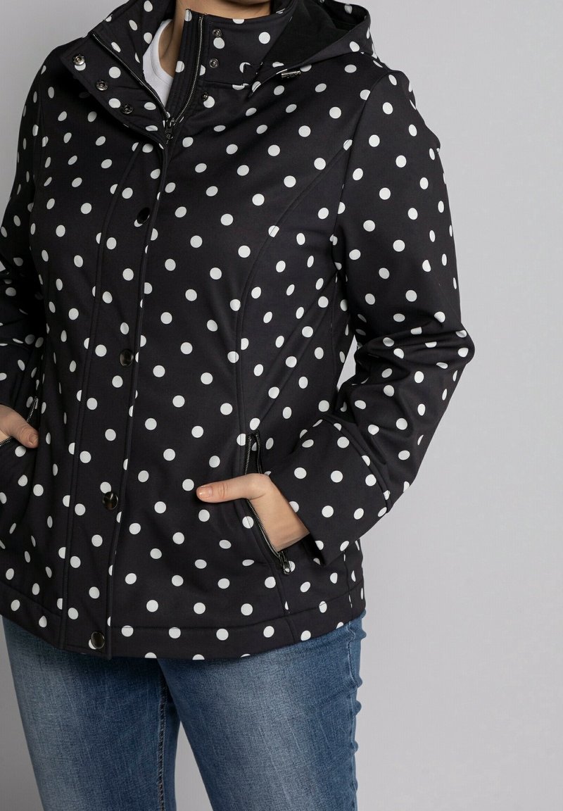 Chaqueta negra impermeable con lunares blancos, que presenta un cierre frontal con cremallera, cierres de botón y bolsillos laterales. Tejido texturizado, diseño con capucha.