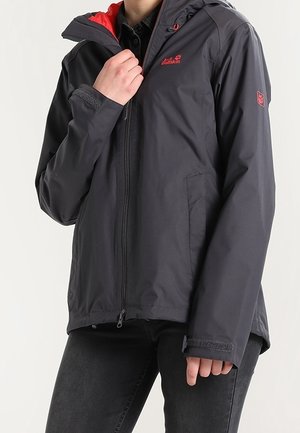 Persona con chaqueta gris oscuro con forro rojo y logo, ajustando la cremallera frontal, combinada con pantalones negros.