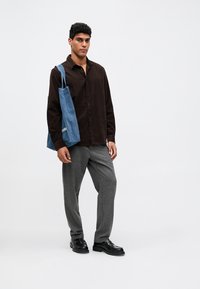 Młody mężczyzna stoi w ciemnobrązowej koszuli z guzikami, szarych spodniach, czarnych butach, niosąc niebieską torbę denimową na ramieniu.