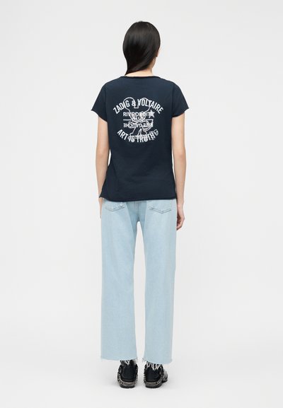 Γυναίκα που φοράει μαύρο T-shirt με λευκό κείμενο και σχέδιο στην πλάτη, συνδυασμένο με ανοιχτό μπλε παντελόνι φαρδύ και μαύρα αθλητικά παπούτσια.