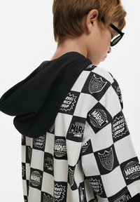 Svart och vit randig hoodie med Marvel-logotyper och grafik. Tillverkad av bomull, lös passform, med en kontrasterande svart huva.