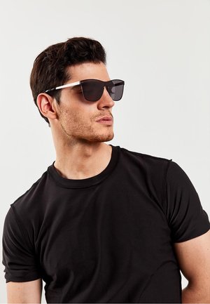 Black sunglasses with a sleek, geometric design and dark lenses. White accent on the temple. Worn by a model in a black T-shirt.