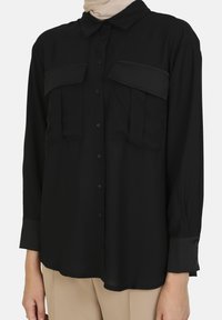 Black button-up shirt made of smooth fabric, featuring two chest pockets, long sleeves with buttoned cuffs, and a classic collar design.
