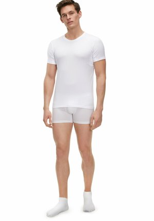 T-shirt blanc ajusté et boxer semi-transparent blanc, tous deux en tissu lisse, associés à des chaussettes blanches, se tenant sur un fond uni.
