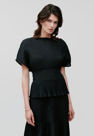Black pleated dress with short sleeves and a fitted waist. Subtle sheen, flowing skirt, and a layered peplum detail at the hem.