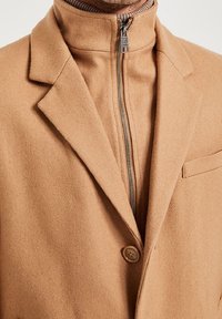 Ljusbrun ullblandad kappa med hög krage, dragkedjestängning fram, en knapp och texturerad yta. Har en bröstficka och strukturerade slag.