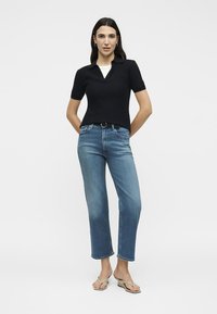 Γυναίκα όρθια με τα χέρια πίσω, φοράει μαύρο πόλο με κοντά μανίκια, μπλε τζιν με ζώνη και μπεζ πέδιλα με τακούνι σε λευκό φόντο.