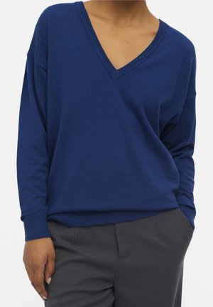 Person iført en løstsiddende blå V-hals sweater og mørkegrå bukser, stående med den ene hånd i lommen mod en ensfarvet baggrund.