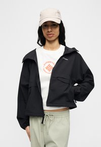 Νεαρή γυναίκα που φοράει μπεζ καπέλο, μαύρο μπουφάν με φερμουάρ, λευκή μπλούζα με το λογότυπο Columbia και ανοιχτό μπεζ παντελόνι με κορδόνι στη μέση, στέκεται μπροστά σε ένα απλό φόντο.