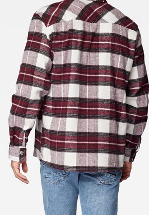 Mężczyzna ubrany w czerwoną, czarną i białą koszulę w kratę z długim rękawem oraz jasnoniebieskie dżinsy, widziany od tyłu na jednolitym tle.