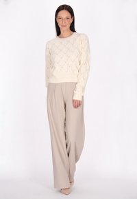 Maglione lavorato a maglia crema con un motivo a diamante, abbinato a pantaloni palazzo beige chiaro. Il modello è in piedi contro uno sfondo neutro.