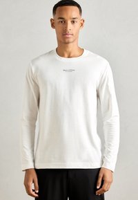 Tânăr îmbrăcat cu o cămașă albă cu mâneci lungi, având un mic logo "Marc O'Polo" pe piept și pantaloni negri, stând în fața unui fundal uniform de culoare deschisă.