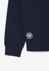 Marinblå sweatshirt med texturerad tyg, ribbade muddar och en cirkulär logotyp med texten "ROLAND GARROS PARIS" som accent.