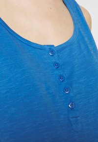 Modrý bezramenný top s kulatým výstřihem, který má vpředu šest modrých knoflíků, vyrobený z lehké, mírně texturované tkaniny.