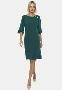 Sukienka do kolan w ciemnozielonym kolorze, o prostym kroju, z falbaniastymi rękawami i dwoma dekoracyjnymi guzikami na lewym ramieniu. Gładka tekstura materiału.