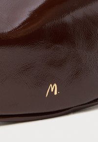Blizgantis tamsiai rudas odinis batų pirštas su mažuoju auksiniu "M." logotipu šalia krašto ant baltos fono.