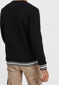 Černá mikina s dlouhými rukávy a kulatým výstřihem, se striped lemem v bílé barvě. Textura vypadá hladce, s uvolněným střihem.
