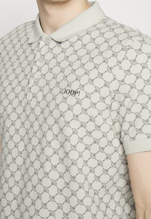 Homme portant un polo beige à motif géométrique avec le logo "JOOP!" brodé sur la poitrine, manches courtes visibles.