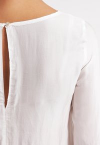 Blusa bianca in tessuto texturizzato. Presenta un'apertura a V sul retro del collo e una piccola chiusura con bottone nella parte superiore.