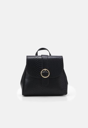 Kleiner schwarzer Rucksack mit strukturierter Oberfläche, Klappenverschluss, goldener runder Schnalle und obenliegendem Griff vor einem schlichten hellen Hintergrund.