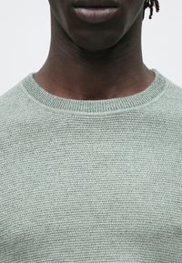 Közeli kép egy személy alsó arcáról, nyakáról és felső mellkasáról, aki világosszürke kötött pulóvert visel, egyszínű háttér előtt.