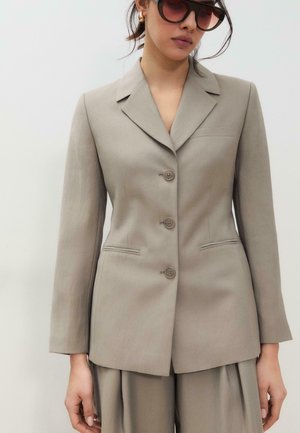 Kobieta w beżowej, dopasowanej marynarce na trzy guziki z wycięciem na klapach oraz luźnych spodniach w tym samym kolorze, uzupełniona dużymi, ciemnymi okularami przeciwsłonecznymi.