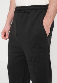 Μαύρο παντελόνι φόρμας με υφή, διαθέτει ελαστική μέση και διακριτικές κεντητές λεπτομέρειες με σταυρό στο πλάι.