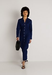 Σακάκι από τζιν σε σκούρο μπλε χρώμα με γιακά, μακριά μανίκια και κουμπιά μπροστά; μοντέλο κρατάει μια μαύρη τσάντα υφής.