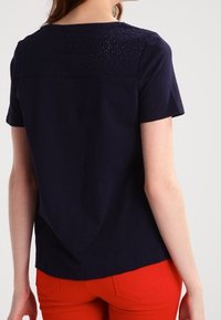 Marinblå kortärmad topp med spetsdetaljer på oket, kombinerad med röda byxor. Slät textur, avslappnad passform och rund halsringning.