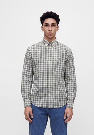 Junger Mann mit lockigem Haar, der ein grau-schwarz kariertes Hemd mit Knöpfen und blaue Jeans trägt, steht vor einem schlichten weißen Hintergrund.