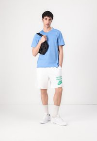 Mėlyna trumpa rankovė marška, balti šortai su žaliu Nike logotipu, juoda per petį nešiojama krepšys, balti sportbačiai, tekstūruota medžiaga ir ilgio iki blauzdos kojinės.