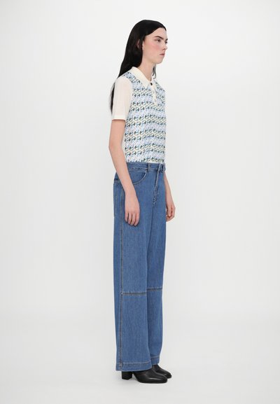 Adīta bezpiedurkņu blouse ar daudzkrāsainu rakstu, krēmkrāsas apkakle; saskaņota ar platajām ziliem džinsu biksēm un melnā augstpapēžu kurpēm. Sānu skats.