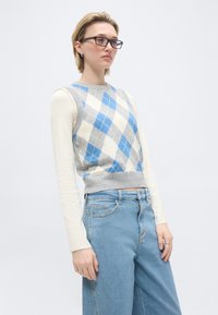 Άτομο με γυαλιά, που φοράει μπλε και γκρι πουλοβεράκι με μοτίβο αργκλάι το οποίο καλύπτει ένα εκρού μακρυμάνικο μπλουζάκι, και ανοιχτό μπλε ψηλόμεσο τζιν, που στέκεται μπροστά σε λευκό φόντο.