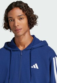 Navy blue zip-up hoodie in soft fabric, featuring a drawstring hood, white striped accents on sleeves, and a white logo on the chest.