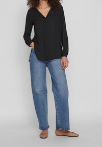 Maglietta nera a maniche lunghe con design a V, abbinata a jeans a gamba larga azzurri e ballerine marroni. La maglietta ha una trama liscia.
