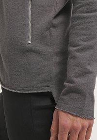 Gros plan sur une personne portant un haut à manches longues gris avec une fermeture éclair à l'avant et un pantalon noir, montrant la partie inférieure de la manche et la main.