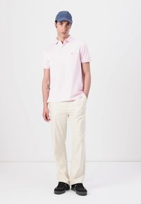 Lichtroze polo met korte mouwen, gecombineerd met crèmekleurige broeken en zwarte sneakers. Het model draagt een blauwe pet.