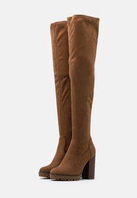 Brązowe zamszowe kozaki za kolano na masywnym obcasie, z gładkim wykończeniem i zaokrąglonym noskiem. Podeszwa z gumy zapewniająca przyczepność.
