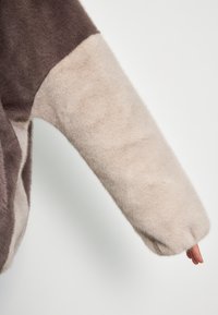 Indumento a maniche lunghe in pelliccia sintetica caratterizzato da un design a blocchi di colore in marrone scuro e beige, con una finitura morbida e testurizzata sulle maniche.