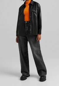 Černá košile z umělé kůže oversized střihu, oranžový žebrovaný top s vysokým krkem a volné černé džíny s viditelnými předními kapsami. Masivní černé platformové boty.