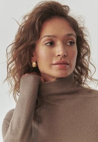 Zlatne naušnice u obliku suze, nosi bež turtleneck pulover. Teksturirana kosa, meki ton kože, prirodni make-up, minimalistička pozadina.