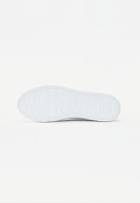 Valge sneakerite tallal on tekstureeritud kumm, mille pinnal on korduv mustriga trükitud brändi nimi "GUESS".