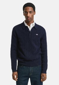 Jonge man in een marineblauwe kabeltrui over een witte met kraag shirt, staande tegen een effen lichte achtergrond.