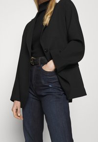 Svart kavaj med vida ärmar och en enda knapp, bärs över en svart polotröja. Kombination med mörka jeans och ett strukturerat svart bälte.