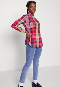 Camisa xadrez vermelha e verde com mangas longas, dois bolsos no peito e um hem em curva. Usada com jeans azul claro e sapatos pretos.