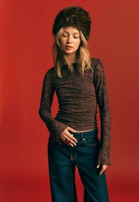 Smeđa long-sleeve majica s reljefnim cvjetnim uzorkom, uparena s tamnim trapericama visokog struka i krznatom kapom, na crvenoj pozadini.